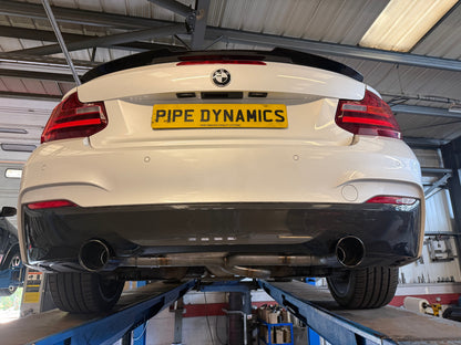 Rear view of a white BMW 2 Series coupe on a blue automotive lift in a workshop, showing a custom black rear diffuser and exhaust system FROM PIPE DYNAMICS